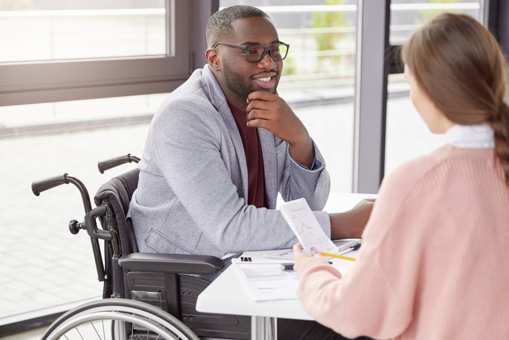 Middle aged dark skinned male invalid sits in wheelchair, works at office, looks with pleased expression at beautiful female assistant, listens to financial report. Job, people and disability concept